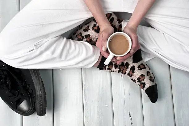 Teenage girl sitting on floor holding a cup of coffee. Body part close up on wooden surface