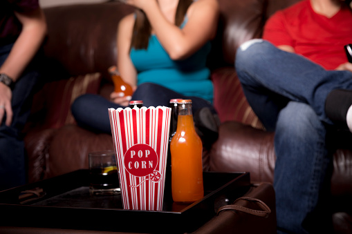 Three young adult friends or siblings hang out and watch TV at home while eating popcorn and drinking sodas. Latin male and female.