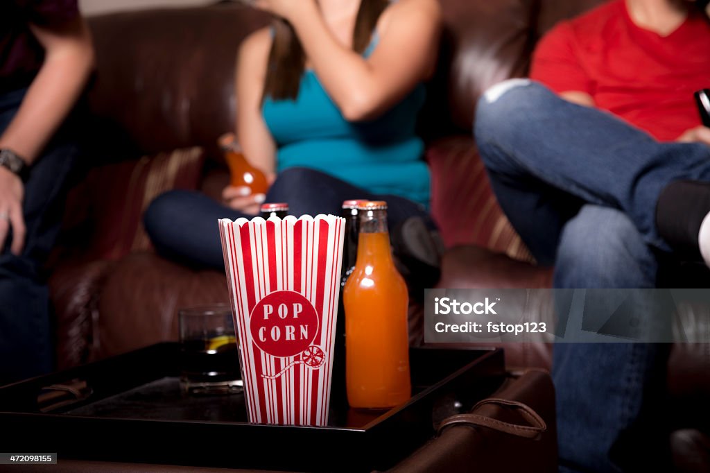 Food and Drink: Young adults hang out. Popcorn, sodas. Three young adult friends or siblings hang out and watch TV at home while eating popcorn and drinking sodas. Latin male and female. Orange Soda Stock Photo