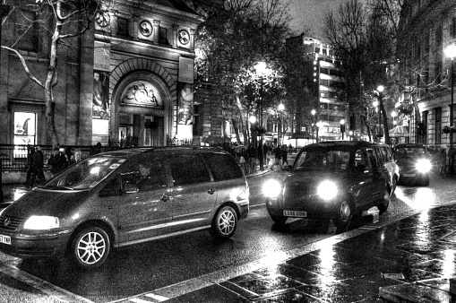 London, United Kingdom - December 15, 2013: London street scene in pouring rain with a people carrier and a blsck cab with headlights on prominent in the picture. There are a large number of tourists shoppers sheltering under umbrellas and hoods walking along this street near Trafalgar Square. The image was shot with a high ISO and post processed in HDR software as well as Photoshop to reduce noise.
