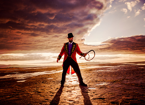 Circus Ring Master in a Dramatic Desert Setting. He is standing with a sun setting the background, which is casting a shadow toward the camera. He is also holding a whip. Full length, square, color image.