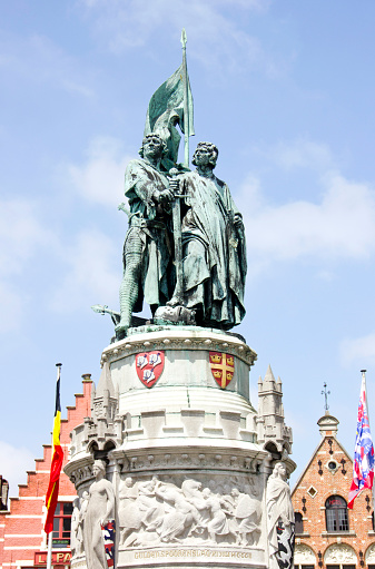 Bruges, Belgium - June 4, 2013: Statue in the middle of picturesque Bruges, Belgium, with colorful flags and buildings in background. Statue of Pieter De Coninck at Market Square in Bruges, Belgium. 