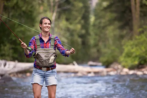 A woman fly fishing for trout on a river in the western United States. She is smiling and casting her line while wading near a shallow riffle.