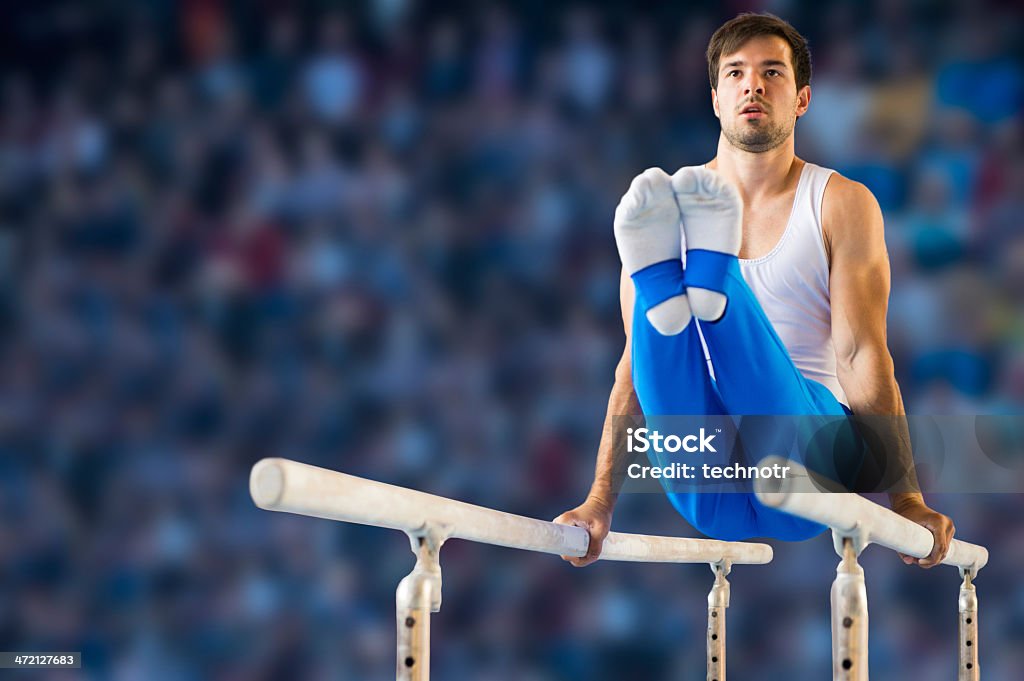 Male gymnast performing routine on parallel bars A young male gymnast performs a routine on the parallel bars.  He balances himself with only his straightened arms on the bars while his legs are held perpendicular to his body.  A crowd of spectators is blurred in the background. Artistic Gymnastics Stock Photo