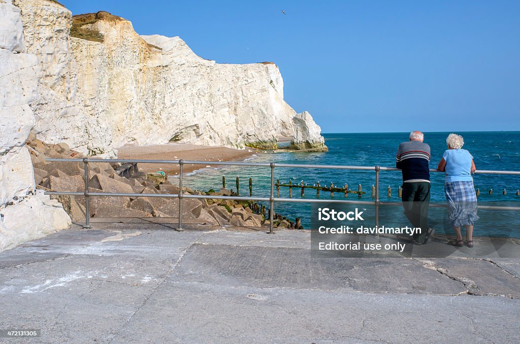 sussex Seaford, UK - July 12, 2013: This is the East Sussex coastal seaside resort of Seaford. This is sea front and beach and the white chalk cliffs of Seaford head are in the background. There are tourist, visitors and local residents relaxing on the shingle beach in the summer sunshine. It is a warm sunny July day in the middle of a summer heatwave. There are two seniors in the foreground looking out to sea. Beach Stock Photo