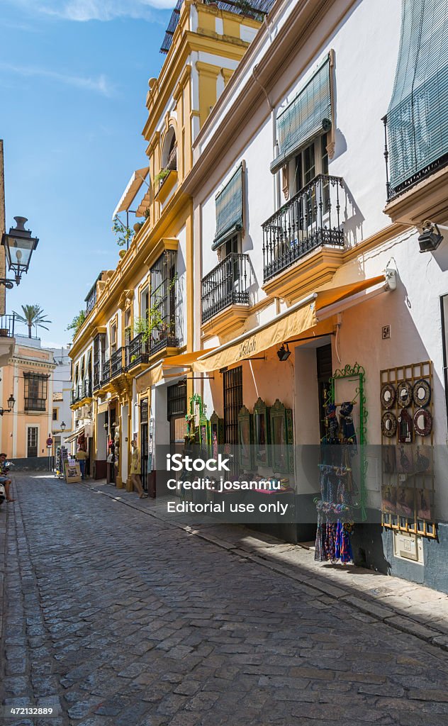 Typical street Sevilla, Andalucia, Spain - September 8, 2013: Typical Street. Typical street in the Barrio de Santa Cruz, Seville. We can see, on the right, the houses with their balconies and in the lower floors, craft shops and souvenirs for tourists. In the background, in the last shop, two seniors, contemplate the goods on display. On the left, at the end of the street, a man takes a beer sitting on his scooter. Adult Stock Photo