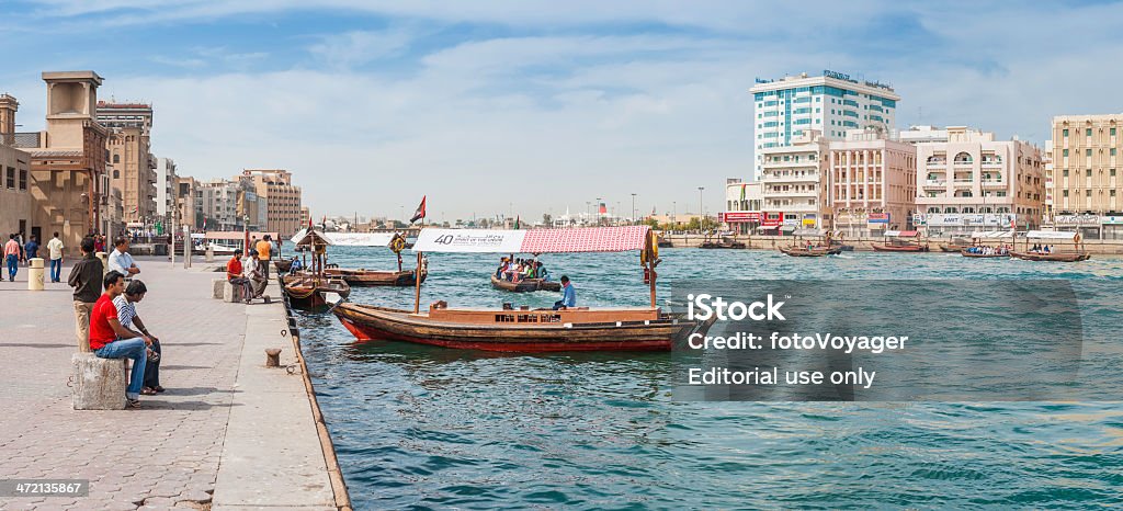 Dubai Creek people relaxing beside busy waterway cityscape panorama UAE Dubai, UAE - 10th February 2012: Locals and immigrant workers enjoying their Friday break on the crowded waterfront of Dubai Creek as traditional abra ferries ply their trade across the river to Deira. Composite panoramic image created from six contemporaneous sequential photographs. Arabia Stock Photo