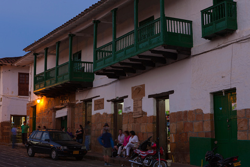 Barichara, Colombia - January 13, 2014: The street on the East side of the town square in the Andean town of Barichara in the department of Santander in the South American country of Colombia.  The town is famous for its Spanish colonial architecture.  It is as if time has stood still here for the last 300 years: the buildings and streets are very well preserved.  Some people both local and tourists can be seen seated on the sidewalk outside a shop, having a drink; above them are some typical balconies.  It is twilight time.