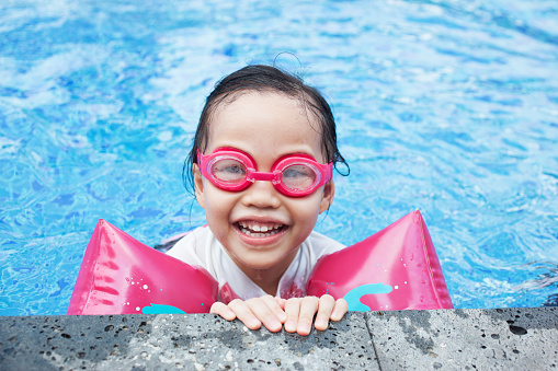 Portrait of a little girl wearing goggles and water wings in a swimming pool, looking at camera with toothy smile.
