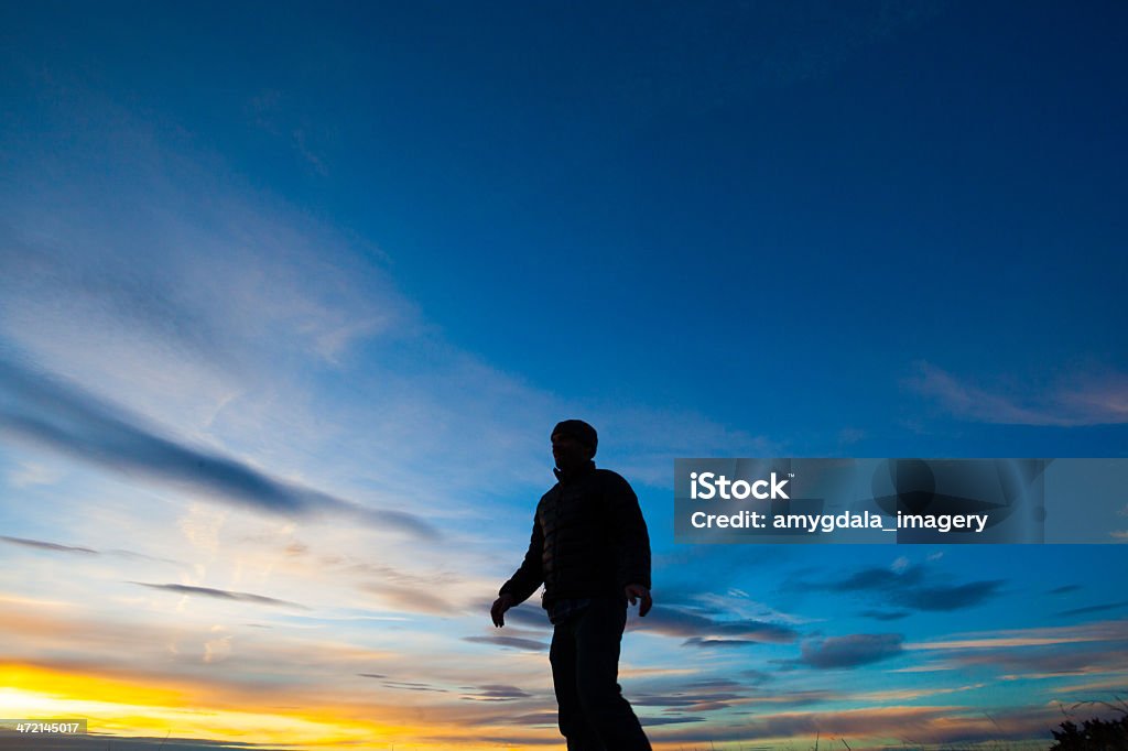 silhouette man watching the sunset sky landscape a silhouetted man stands watching cloud filled blue and yellow sunset sky in the sandia mountains of albuquerque, new mexico. horizontal wide angle composition with copy space. Adult Stock Photo