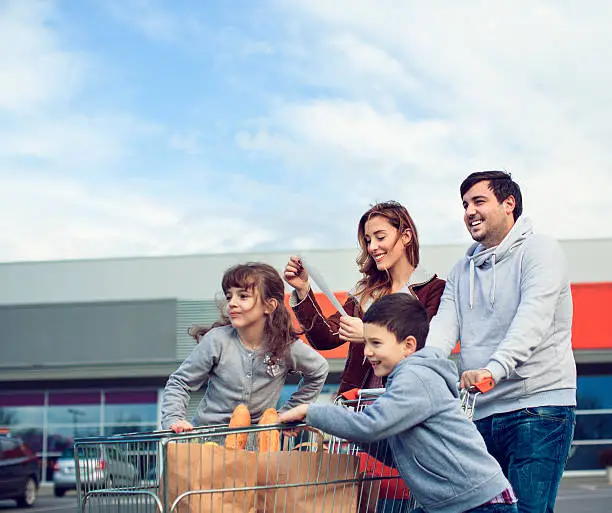 Cheerful family going back from supermarket. Pushing shopping cart and carrying shopping bags. Mother looking at shop bill.