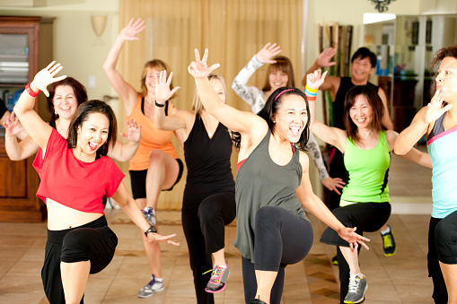 A multi-ethnic group of women raising their arms and dancing together during a workout class in a gym studio wearing exercise clothes.