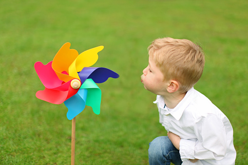 Little boy is blowing a colorful pinwheel toy outdoors in the garden against green grass background. The child is seen from the side, while the windmill is seen from the front.