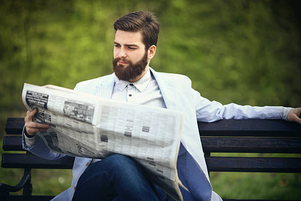 Front portrait of handsome 30's bearded caucasian man reading newspapers in local park.He has full grown beard and neat brown hair.His legs are crossed,wearing smart casual jacket and dark blue jeans. Blurry grass in background.