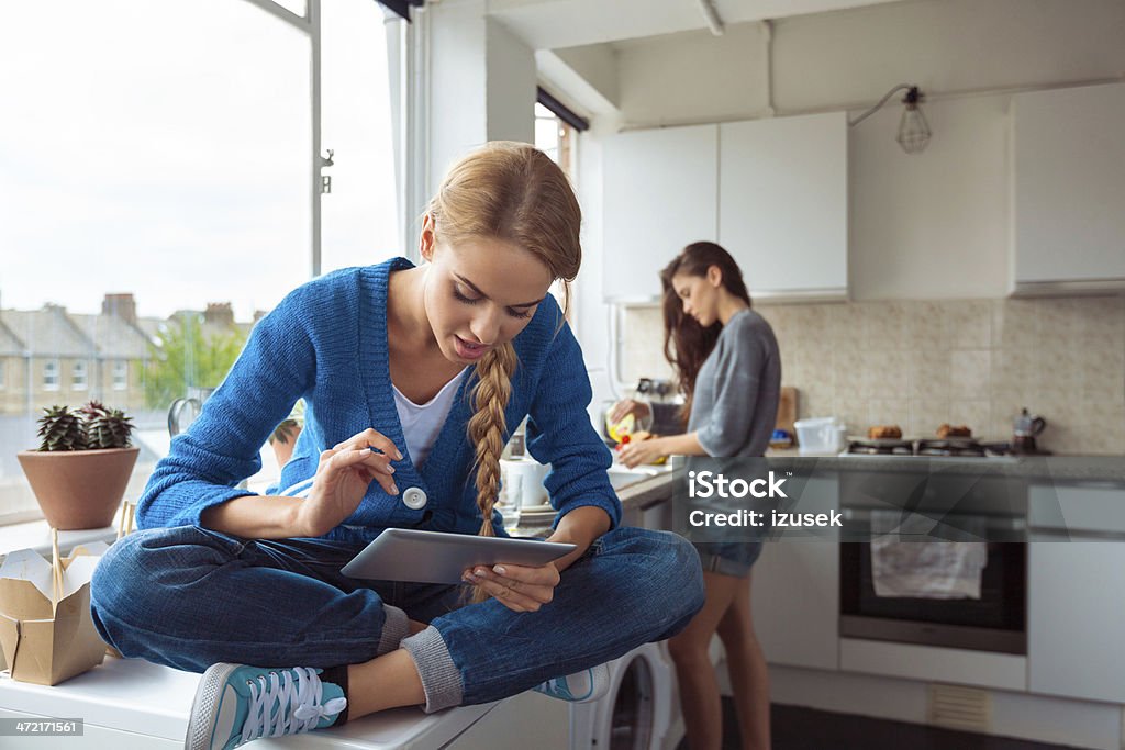 Girl using digital tablet Roommates lifestyle. Young woman using a digital table while her friend washing dishes in the background. Student Stock Photo