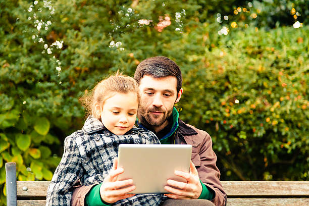istock Father and daughter sitting on bench looking at a tablet. 472172099