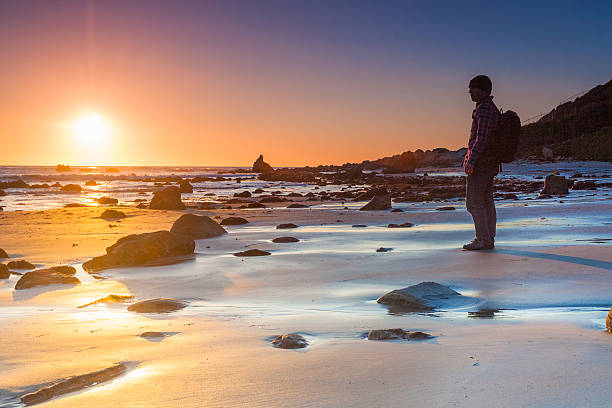 silhouette man beach sunset landscape a man stands watching the sun reach the horizon at sunset along a boulder strewn coastline with the pacific ocean.  such beautiful nature and outdoor peace of mind can be found at el matador beach in malibu, california.  horizontal wide angle composition.   rock sea malibu silhouette stock pictures, royalty-free photos & images