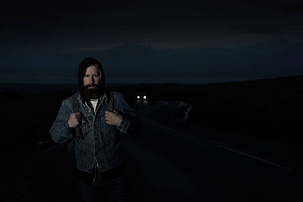 Mid 30's Bearded Casually Dressed man with backpack walking along a twisty country road during dusk, Car with lights approaching to his rear.