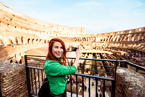 happy tourist at colloseum making photo with her smart phone, casual wear, red hair and happy expression.