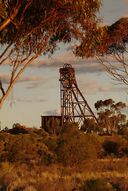 An old mining headframe used to transport workers and equipment down a mineshaft. The headframe is located in woodlands and is seen at a distance through trees at sunset. Vertical photo.