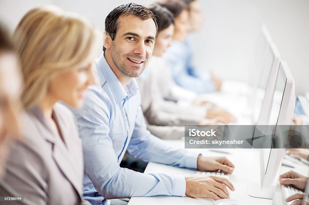 Business people on a computer class. Group of business people sitting in a row in a computer lab. Focus is on smiling mid adult man looking at the camera.    Business Stock Photo