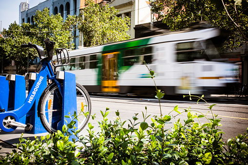 A tram passing by in city center of Melbourne in Australia on a sunny day. The tram is blurred due to motion and long exposure. A Melbourne Bike Share system is in the foreground.