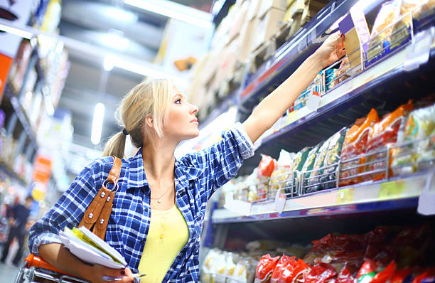 Woman buying food in supermarket. Young blond woman buying frozen food in local supermarket.She's reaching for a product on the shelf.She's looking for pasta. Pasta aisle stretch blurry behind her.She has light blond hair pulled back with a clip and wearing blue shirt..She's holding shopping list in other hand. convenience food photos stock pictures, royalty-free photos & images