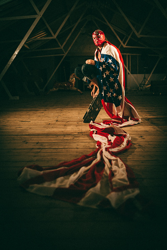 mexican american luchador, young guy wearing a lucha libre mask and a huge usa flag appears to be waiting for its next match inside a weird warehouse.