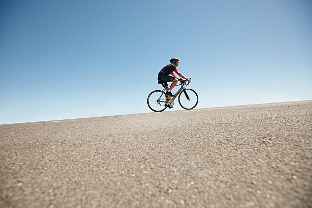 ciclista masculina dirigindo em uma estrada plana contra o céu azul - colina acima imagens e fotografias de stock