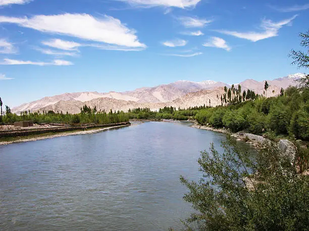 Himalayan river Sindhu, at Leh, Ladakh, India. At Leh, the Sindhu river is not merely a rivulet. At leg, the river look wide and deep and fast flowing blue waters. The himalayan mountains are visible at the horizon. Both the banks of the river are surrounded by green trees and foliage.
