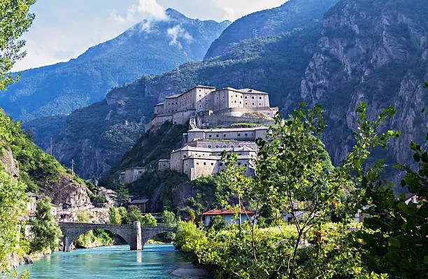 Overall view of the Fort of Bard (Aosta, Italy) from the banks of the River Dora Baltea, which is crossed by an ancient stone bridge. In the background can see the mountain range of the Alps and the foothills of the fort lies the ancient village. Courtesy Forte di Bard.