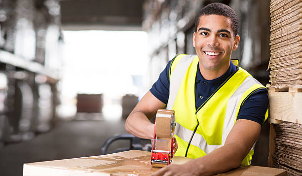 A young male warehouse worker is using a tape dispenser gun to secure a cardboard box shipment in a large distribution warehouse. he is wearing a hi vis jacket .