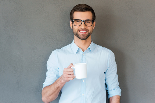 Handsome young man in shirt in eyewear holding cup of coffee and smiling at camera while standing against grey background