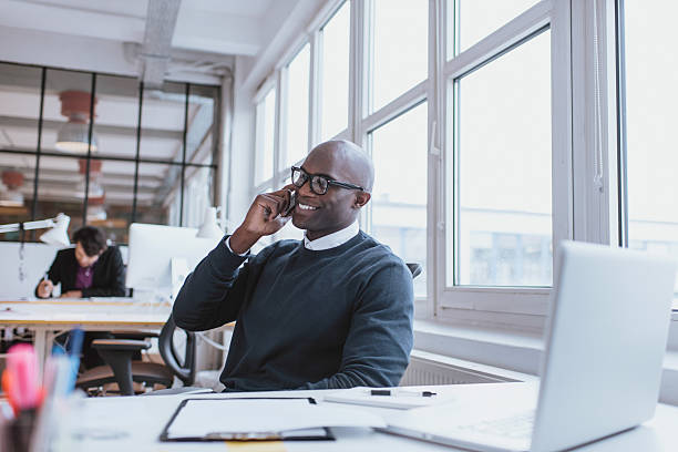 Young man talking on his mobile phone in office Young man talking on his mobile phone in office. African executive sitting at his desk with laptop small office stock pictures, royalty-free photos & images