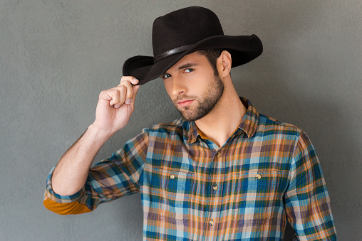 Handsome young man adjusting his cowboy hat and looking at camera while standing against grey background