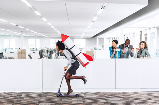 Woman speeding thru the office on the scooter with a rocket strapped to her back while her happy multiethnic  co-workers are watching her succeed in the workplace. Plenty of copyspace. 