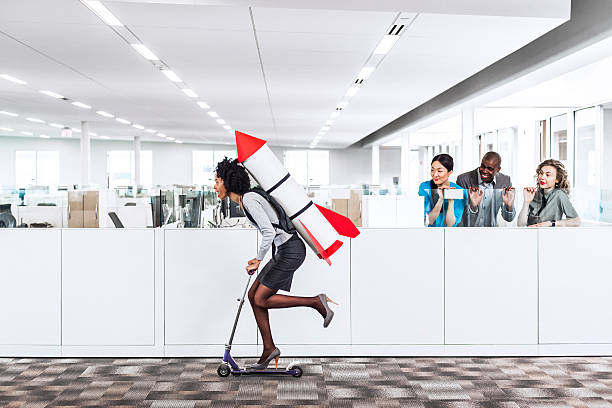 Moving forward Woman speeding thru the office on the scooter with a rocket strapped to her back while her happy multiethnic  co-workers are watching her succeed in the workplace. Plenty of copyspace.  office cubicle photos stock pictures, royalty-free photos & images