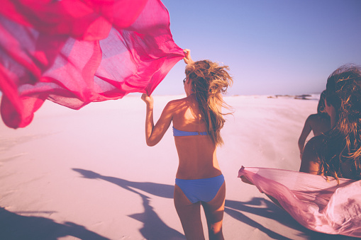 Rearview of a girl in a bikini running on a beach flying a piece of bright pink cloth behind her