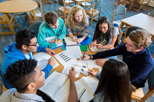 Diverse group of teen high school students are sitting around round table in library. They are studying books and papers to prepare for a test. Caucasian, African American, Hispanic, and Indian teen boys and girls are smiling while working together on class project. 