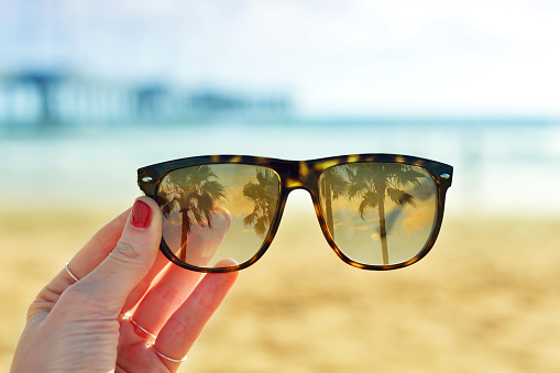 Young woman holding sunglasses on the beach. Palm trees reflection in the glass and beach at the background.