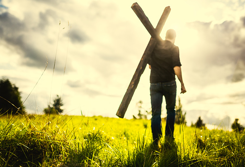 A man walks through a bright and sun lit wilderness area, carrying a large wooden cross over his shoulder.  A depiction of a Christian image given in the Bible, of picking up ones cross and following Jesus Christ.  Horizontal image with copy space in the sky.