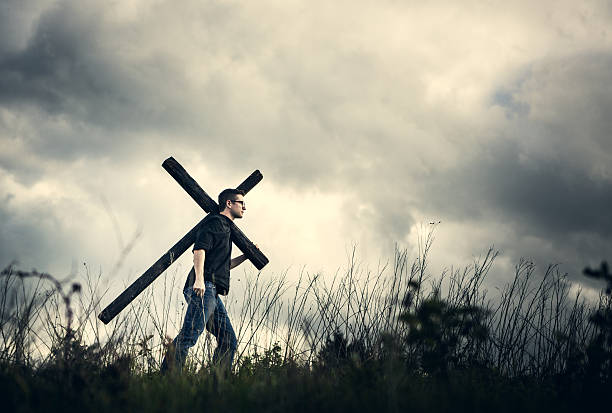 A man walks through a desolate dark wilderness area, carrying a large wooden cross over his shoulder.  A depiction of a Christian image given in the Bible, of picking up ones cross and following Jesus Christ.  Horizontal image with copy space in the clouds.