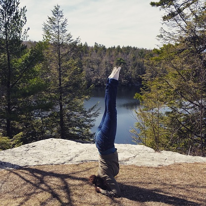 This is a square, color, royalty free mobilestock photograph shot with a Samsung Galaxy S5 on a spring day in New York State's Catskill Mountains. Pine trees line Lake Minnewaska and are reflected in the calm blue water surface. An American woman wearing jeans practices her yoga headstand on a cliff overlooking the scenery.