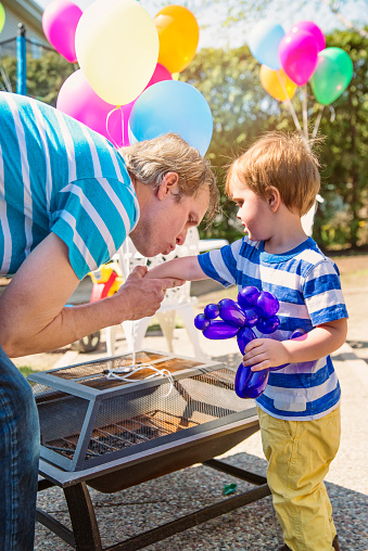 Dad is making his son feel better kissing his hurt arm. Both are wearing striped t-shirts and are at a birthday party, with balloons in the background. Kid is holding a purple balloon doggie. Vertical candid shot on a bright and sunny spring day.