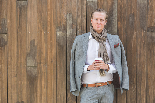 Fashioned young man in Oslo holding a cup of coffee and leaning on a wooden background. He has nordic facial features, and wearing a light gray suit.