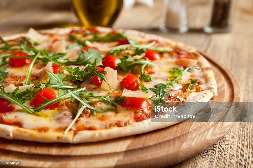 Italian pizza "Caprese" on a wooden table. Italian pizza "Caprese" lies on beautiful wooden table. At the top of a pizza cut, baked cherry tomatoes, arugula and Parmesan cheese slices. Nearby are the containers with spices and olive oil. Pizza Stock Photo