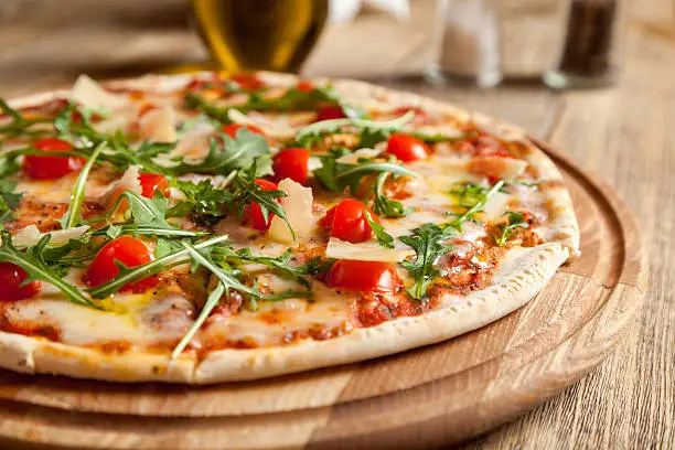 Italian pizza "Caprese" lies on beautiful wooden table. At the top of a pizza cut, baked cherry tomatoes, arugula and Parmesan cheese slices. Nearby are the containers with spices and olive oil.