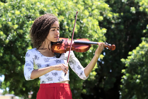 Half length portrait of a young African American woman wearing a white patterned blouse, red skirt, and playing violin.  Woman is outdoors with trees in the background.  Image is landscape orientation.