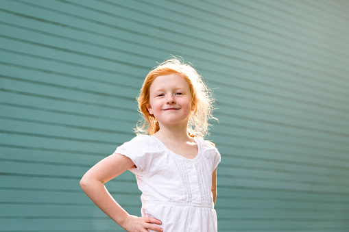 A vibrant and happy young redheaded girl child stands in a strong pose with hands on her hips in a beautiful sun flare against a turquoise wall.