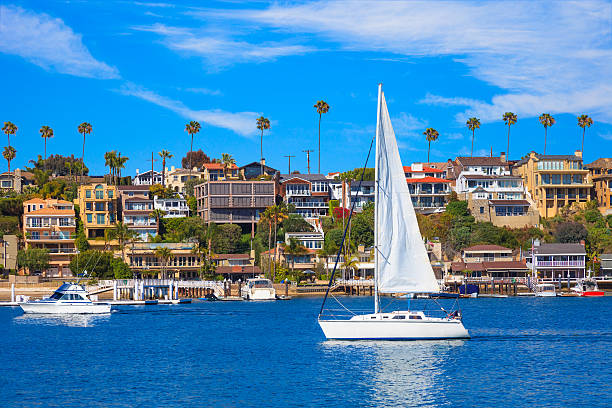 Recreational sailboat on Newport Bay at Newport Beach, CA Recreational sailboat sailing on calm harbor waters of Newport Bay with beach houses and Fashion Island in background. Wispy clouds fill the sky. Newport Beach, California newport beach california stock pictures, royalty-free photos & images